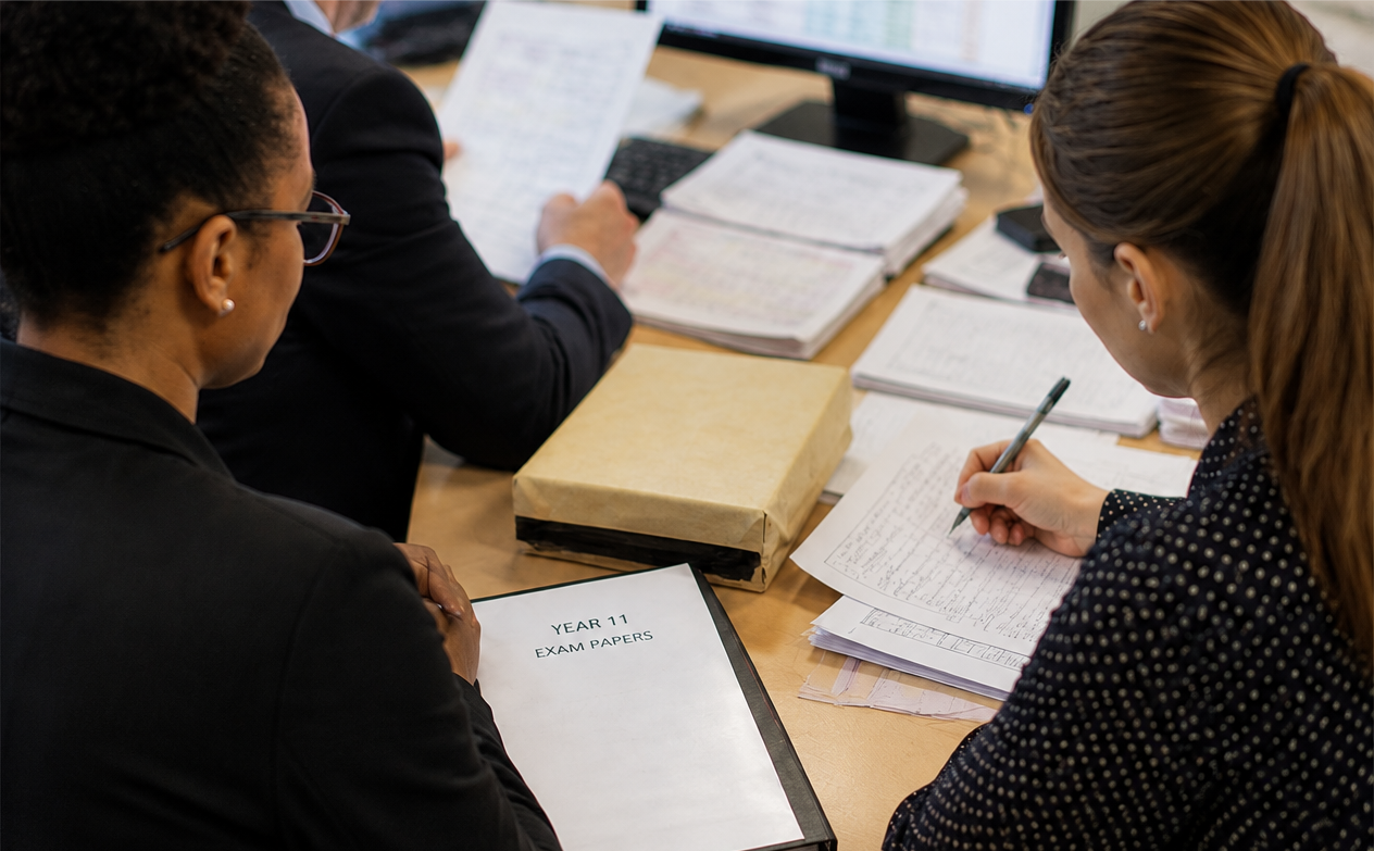 Three people in business attire sit at a desk with documents, one with a folder labeled "Year 11 Exam Papers" and another writing on papers.